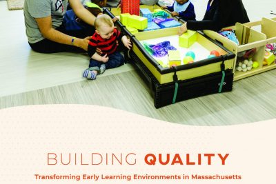 Three adults, three toddlers sitting on the floor playing with building blocks on low tables with bright windows in the background.