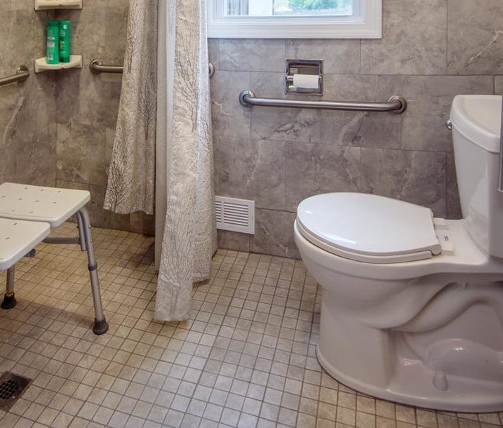 A barrier-free, walk-in shower in a neutral grey tile. The seamless cream color floor tiles extend throughout the bathroom. Additional accessibility features include hand-rails and a shower-chair.